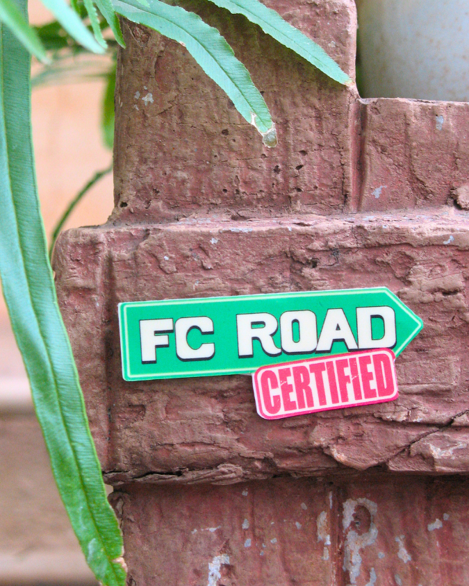 Green and pink 'FC Road Certified' sign on a wooden post with plants in the background